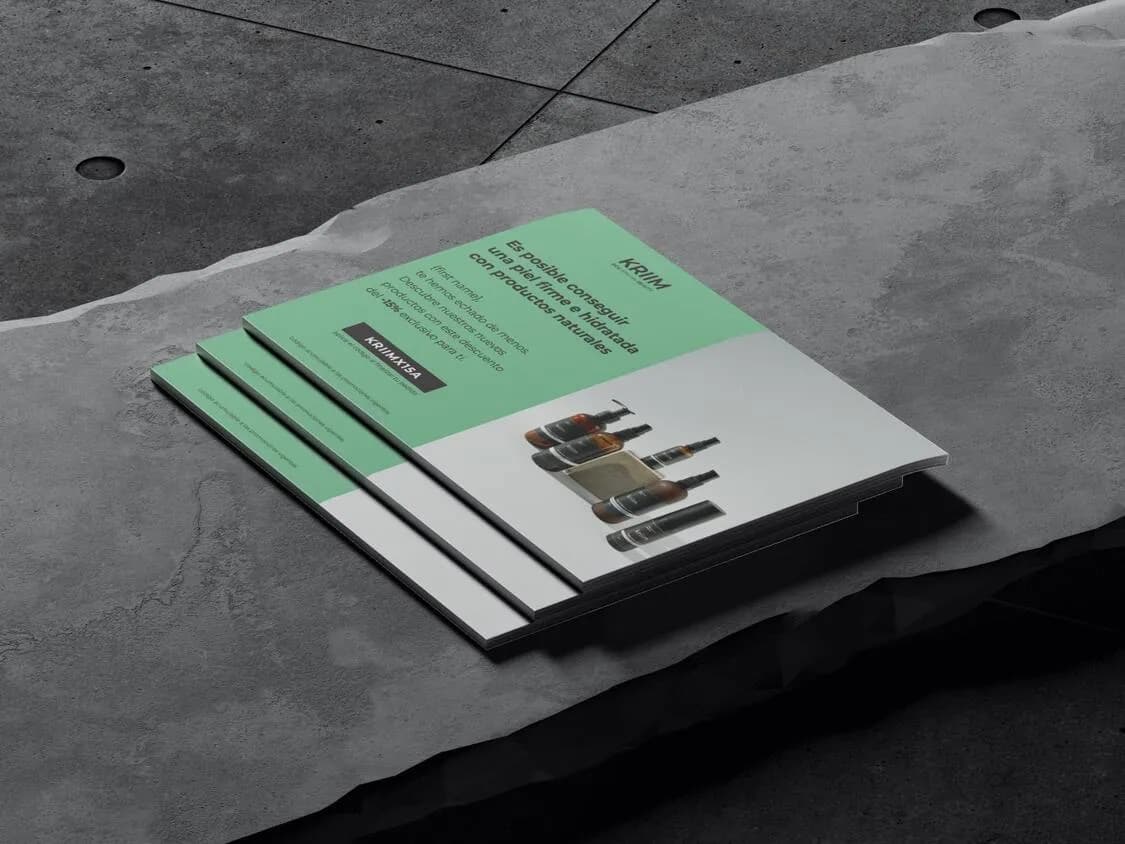 A stack of three printed flyers or brochures with a minimalist design, featuring a mint green header and white body, resting on a textured gray concrete surface with visible seams and circular drain holes in the background; the top flyer displays the brand name 'KRIM' in bold black letters, followed by Spanish promotional text about achieving firm and hydrated skin with natural products, and includes a dark banner with 'KRIMX15A' and images of five skincare product bottles arranged diagonally on the white section; the flyers have a clean, modern aesthetic with sharp edges and slight shadows indicating depth and layering.